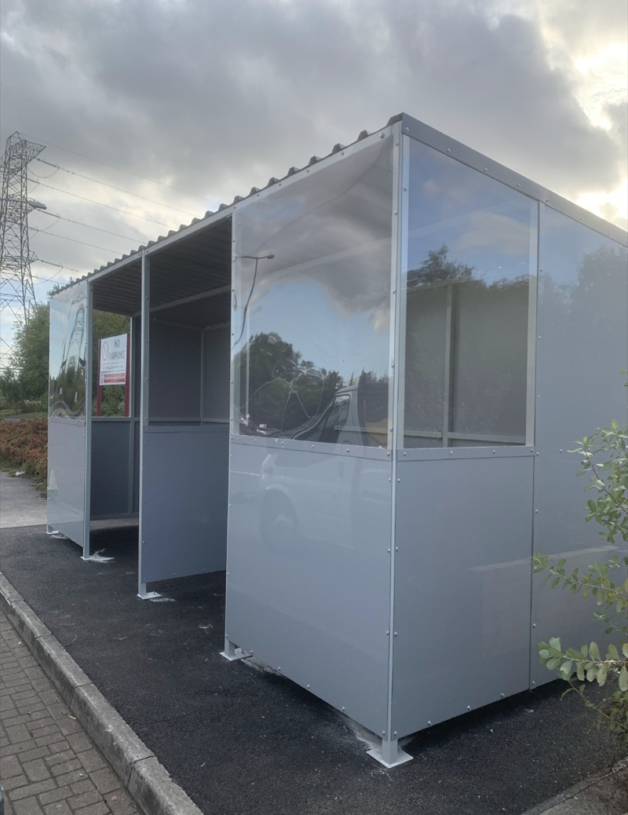 Enclosed grey glazed smoking shelter viewed from the corner on a tarmac bay in Bedford