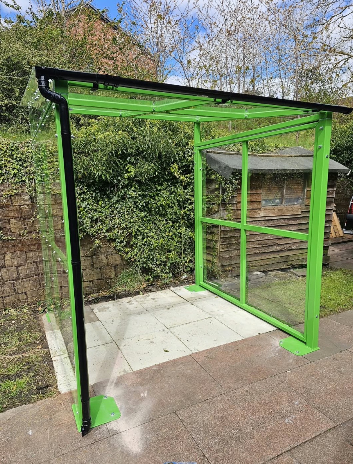 Green-framed glazed smoking shelter on a paved patio beside a hedge in Nottingham